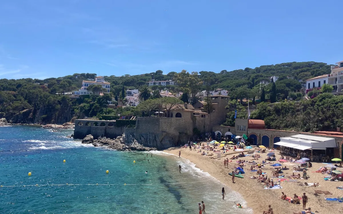 Sandy cove with a stone building and a pine-covered hillside on the Costa Brava