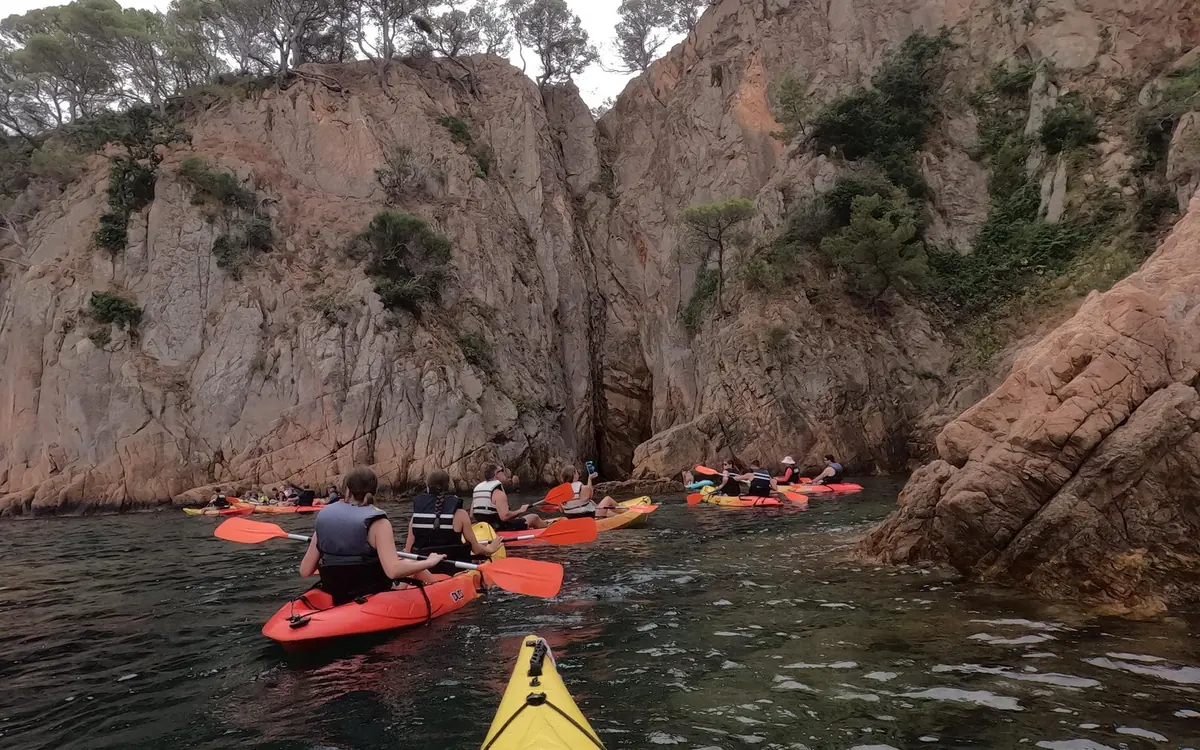 A line of sea kayaks on green water below a split in the Costa Brava cliffs